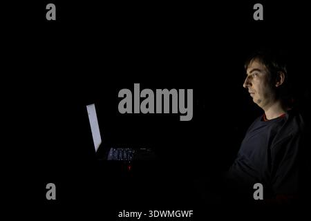 Caucasian man in a dark room lit by his laptop screen. Side view, pitch black room Stock Photo