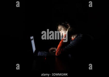 Side profile of an exhausted man resting his head on his hand while looking at a glowing laptop screen in a completely dark room Stock Photo