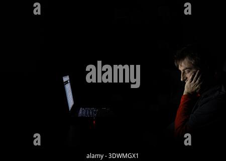 Side profile of an exhausted man resting his head on his hand while looking at a glowing laptop screen in a completely dark room Stock Photo