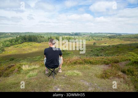 Man sits on stool on hilltop looking out over vast heathland, man and nature, nature therapy and tranquility Stock Photo