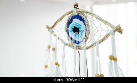 Handmade dreamcatcher amulet with an evil eye nazar woven from natural thread, accented with white feathers and wooden beads, hanging against a bright Stock Photo