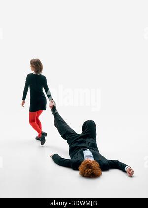 Woman dragging man by leg across studio floor on white background in black formal outfits. Stock Photo