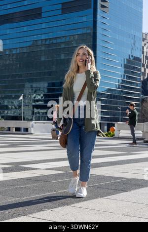 Blonde woman in 20s walking across plaza by glass tower in green jacket, holding phone, skateboard Stock Photo