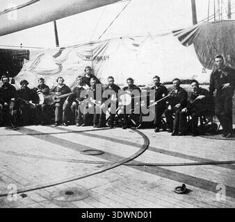 This vintage circa 1861 photograph captures a group of sailors, likely from the USS Wabash, performing as a minstrel troupe on the ship's deck during the American Civil War. The group is performing in front of a painted backdrop featuring the ship's name and "Hatteras", indicating their participation in the Battle of Hatteras Inlet. Stock Photo