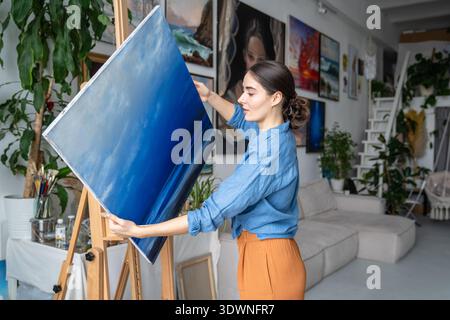 Professional female artist painter carefully taking off canvas from easel. holding finished picture. Stock Photo