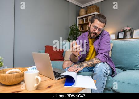 Freelance AI consultant working from home on sofa, smiling during phone call while reviewing analytics reports on laptop, remote technology advisory a Stock Photo