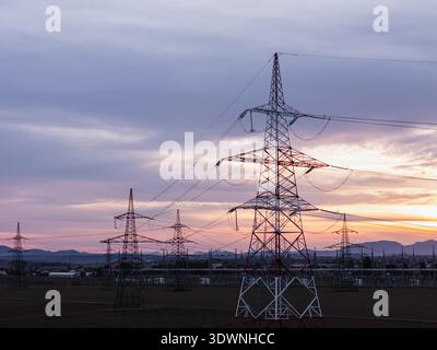 Power lines and electrical towers stand in a rural area as the sun sets, showcasing energy infrastructure against a dramatic sky. Stock Photo