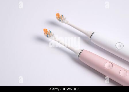 A detailed high-angle shot of two modern sonic toothbrushes, one in pastel pink and one in classic white, lying on a flat white surface. The focus is Stock Photo