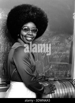 Dr. Valerie Thomas, an African American scientist, is captured in this 1979 black and white photograph, smiling brightly with a large afro and round glasses. She stands beside a stack of Landsat Computer Compatible Tapes (CCTs), with mathematical formulas and technical terms visible on a blackboard behind her, reflecting her pioneering work in digital media formatting for early Landsat data. Stock Photo