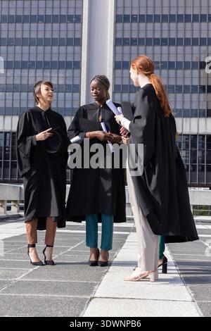 Diverse female graduates standing on roof terrace holding black gowns, diplomas, cap, folders Stock Photo