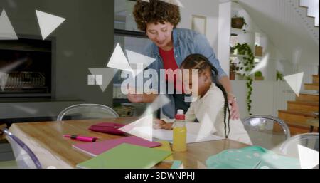 Leaning adult woman wearing denim shirt guiding girl writing at home dining table, with pink tablet Stock Photo