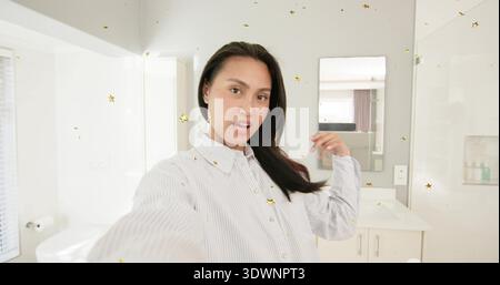 Taking Asian woman in striped shirt adjusting hair with phone, white bathroom vanity and gold stars Stock Photo
