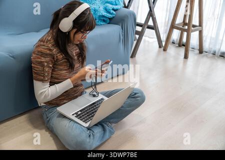 Young woman sitting on the floor at home using a laptop and smartphone while wearing wireless headphones. Concept of remote learning, online education Stock Photo