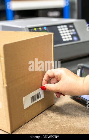 Warehouse operative applying a barcode label onto a cardboard box, with a barcode scanner device visible in the background. Stock Photo