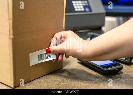 Warehouse operative applying a barcode label onto a cardboard box, with a barcode scanner device visible in the background. Stock Photo