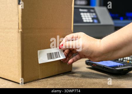Warehouse operative applying a barcode label onto a cardboard box, with a barcode scanner device visible in the background. Stock Photo