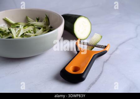 Preparation of zucchini noodles with a professional peeler and a bowl of shredded squash on a light surface Stock Photo
