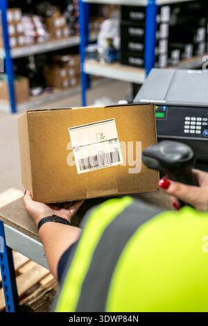 Handheld barcode scanner in use at a retail point-of-sale counter, scanning a product with a label printer and storage racking in the background. Stock Photo