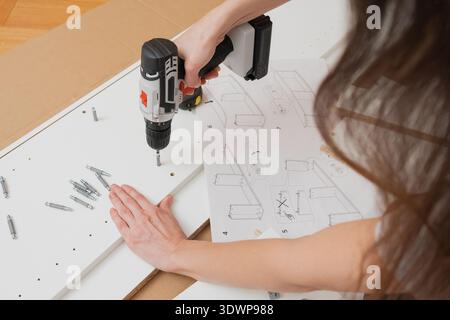 A woman uses a screwdriver to fasten a furniture panel on the floor surrounded by instructions, parts, and tools in home interior Stock Photo