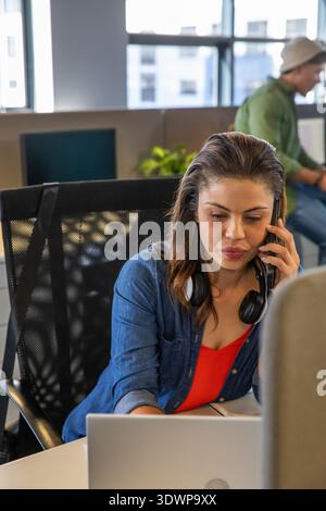Female worker sitting at desk using laptop and talking on smartphone with headphones in office Stock Photo
