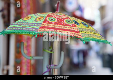 Vibrant hand-embroidered parasol in dazzling green, yellow, and red patterns with fringe and eye motifs hangs from a pole in a colorful shop in Nice Stock Photo