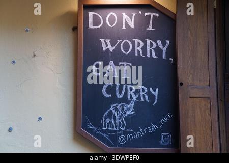 Chalkboard sign at Mantrah in Nice reads "DON'T WORRY, EAT CURRY" with a cheerful elephant drawing, hanging outside an Indian restaurant in the town Stock Photo