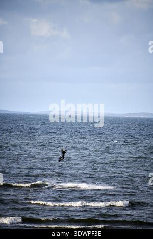 An adventurous male kitesurfer is captured mid-air, performing an impressive jump over the dark, choppy waves of the ocean. Dressed in a wetsuit and h Stock Photo