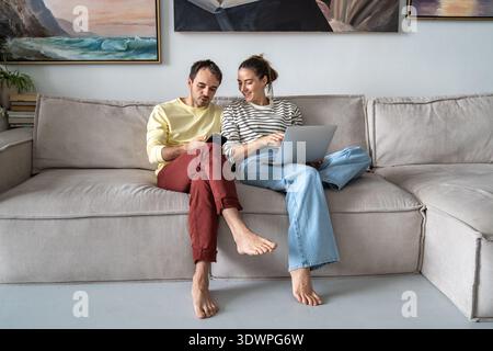 Positive married couple husband and wife relaxing on sofa with smartphone and laptop browsing online Stock Photo