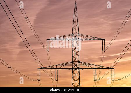 A striking silhouette of a massive lattice steel electricity pylon and power lines against a vibrant, cloudy sunset sky. The golden, pink, and orange Stock Photo