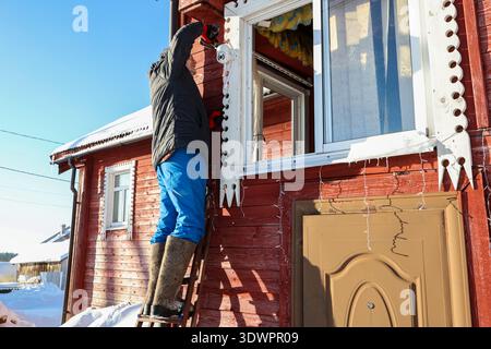 Outdoor surveillance camera mounting on facade of farm house in winter, farmer stands on stepladder holding screwdriver in hands. Stock Photo