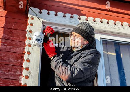 After installing outdoor dome surveillance camera on facade of farmhouse rural resident connects power supply to it Stock Photo