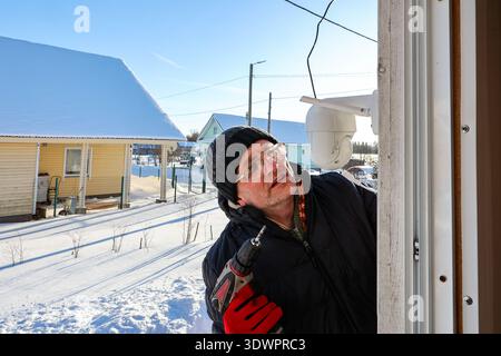 Outdoor dome surveillance camera mounting on facade of rural house, farmer tightens fastener using screwdriver. Stock Photo