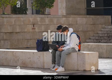 Two male students/friends using a portable computer. Studying or working remotely on a sunlit stone ledge in Valencia, Spain. Stock Photo
