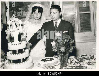 A bride and groom cut their three-tier wedding cake at an indoor reception in Cairo, Egypt, 1960. Flowers and a dessert spread are laid out on the table before them. Stock Photo