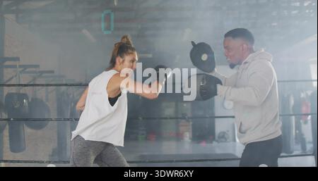 Throwing boxer in white top landing punch with black gloves in boxing ring, trainer holding mitts Stock Photo