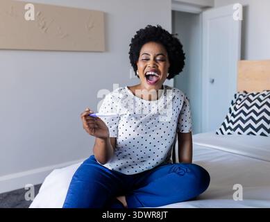African American woman sitting on bed in dot blouse, blue pants, holding pregnancy test, laughing. Softlight, daylight, modern, cozy, polka, lifestyle Stock Photo