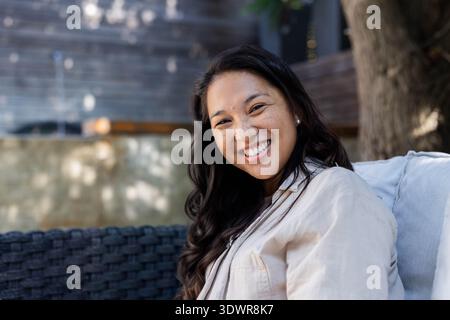 Asian woman sitting on woven sofa in shaded patio, wearing beige shirt, pearl earrings. Outdoor, backyard, casual, relaxation, lounge, bokeh, dappled Stock Photo