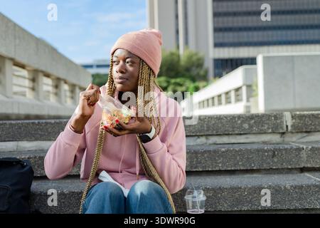 African American woman sitting at campus steps wearing pink hoodie, eating salad from clear box. Urban, outdoors, plaza, casual, student, backpack, lu Stock Photo