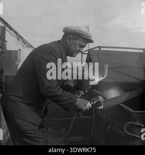 Historical 1970s black and white portrait captures a focused male worker performing welding tasks at the repair workshop of the Donrybkombinat state enterprise in the Slavyansk district, Ukrainian SSR, USSR. Dressed in a checkered suit jacket and a classic cap, he uses a handheld welding shield while working on metal equipment. This authentic archival photograph highlights the essential maintenance of Soviet agricultural machinery, the dignity of skilled industrial labor, and the optimistic atmosphere in the peaceful Donbass before the war. Stock Photo
