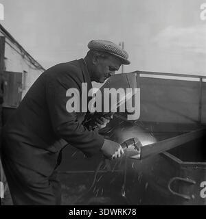 Historical 1970s black and white portrait captures a focused male worker performing welding tasks at the repair workshop of the Donrybkombinat state enterprise in the Slavyansk district, Ukrainian SSR, USSR. Dressed in a checkered suit jacket and a classic cap, he uses a handheld welding shield while working on metal equipment. This authentic archival photograph highlights the essential maintenance of Soviet agricultural machinery, the dignity of skilled industrial labor, and the optimistic atmosphere in the peaceful Donbass before the war. Stock Photo