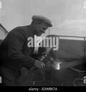 Historical 1970s black and white portrait captures a focused male worker performing welding tasks at the repair workshop of the Donrybkombinat state enterprise in the Slavyansk district, Ukrainian SSR, USSR. Dressed in a checkered suit jacket and a classic cap, he uses a handheld welding shield while working on metal equipment. This authentic archival photograph highlights the essential maintenance of Soviet agricultural machinery, the dignity of skilled industrial labor, and the optimistic atmosphere in the peaceful Donbass before the war. Stock Photo