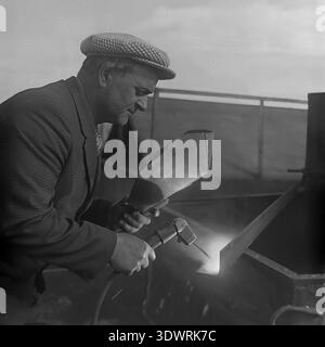 Historical 1970s black and white portrait captures a focused male worker performing welding tasks at the repair workshop of the Donrybkombinat state enterprise in the Slavyansk district, Ukrainian SSR, USSR. Dressed in a checkered suit jacket and a classic cap, he uses a handheld welding shield while working on metal equipment. This authentic archival photograph highlights the essential maintenance of Soviet agricultural machinery, the dignity of skilled industrial labor, and the optimistic atmosphere in the peaceful Donbass before the war. Stock Photo