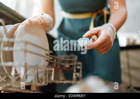 A close-up shot captures a person in gloves placing a dishwasher detergent tablet and loading dishes into a modern dishwasher. The scene evokes a sens Stock Photo