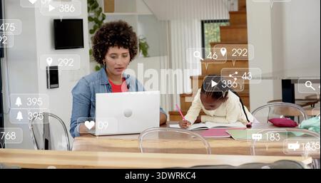 Working woman in denim using white laptop at dining table, child writing with pink pencil, alerts Stock Photo