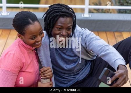 African American couple in athletic wear sitting on deck, taking selfie with smartphone and cup Stock Photo