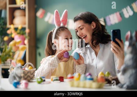 Joyful mother and daughter celebrate Easter at home, taking a selfie together surrounded by colorful painted eggs and festive decorations. The girl we Stock Photo
