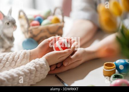 Heartwarming close up of two pairs of hands gently holding a brightly painted Easter egg, surrounded by colorful eggs and springtime elements. The ima Stock Photo