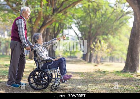 Happy Asian senior man pushing a wheelchair for wife outside in the park, Senior couple and retirement life concept Stock Photo