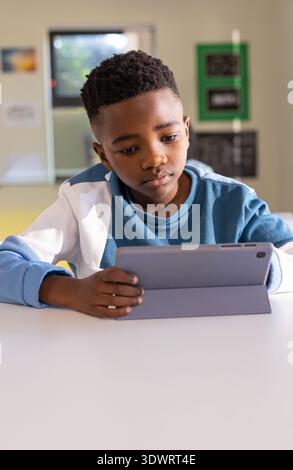 African American child wearing blue crewneck jacket draped sitting in classroom using tablet case. Student, device, touchscreen, education, learning, Stock Photo