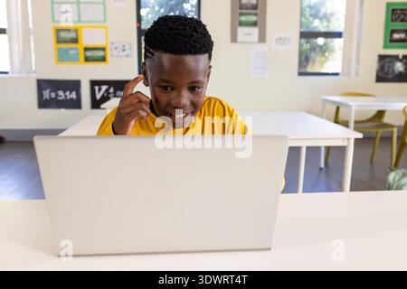 Adolescent African male wearing yellow tee working on silver laptop at school with pi poster. Classroom, student, daylight, desks, chairs, learning, t Stock Photo